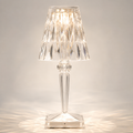 Transparent faceted table lamp glowing on a light studio background with warm reflections around the square base.