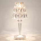 Transparent faceted table lamp glowing on a light studio background with warm reflections around the square base.
