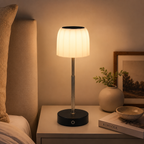 A modern table lamp with a pleated white shade and a black circular base sits on a bedside table in a warmly lit bedroom. The lamp is turned on, casting a soft glow, with a vase, framed picture, and book nearby.