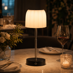 A table lamp with a pleated white fabric shade and a black circular base sits on a dining table, glowing warmly. The scene includes a wine glass, a candle holder, plates, and flowers in a softly lit restaurant or home dining setting.