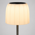 A modern table lamp with a pleated, fluted white shade that glows warm yellow when lit, supported by a slim dark metal stem against a neutral gray background.