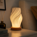A modern wavy LED table lamp with a warm glowing white ribbed shade and a round wooden base, styled on a bedside table next to a reed diffuser and books.