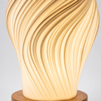 A modern table lamp with a sculptural, fluted spiral design that glows with a warm yellow light, sitting on a round wooden base against a neutral gray background.