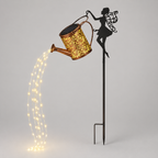 A decorative garden stake featuring a black fairy silhouette holding a glowing copper-toned watering can. The watering can has an ornate cutout pattern and casts warm light downward, simulating water, with string lights visible against a plain light gray background.