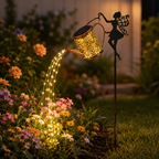 A garden stake featuring a fairy silhouette holding a glowing copper-colored watering can. Warm white LED lights are arranged to follow the spout of the watering can, illuminating flowers and a stone patio in an outdoor garden setting.