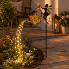 A garden stake featuring a fairy silhouette holding a glowing copper-colored watering can. Warm white LED lights are arranged to follow the spout of the watering can, illuminating flowers and a stone patio in an outdoor garden setting.