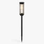 A modern black metal solar landscape light with a glass cylinder shade and a long stake, shown lit against a plain white background.