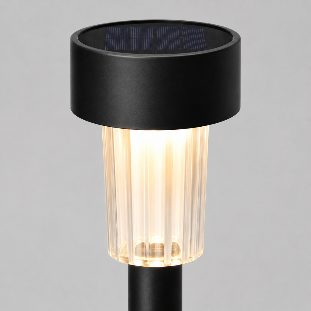 A modern solar-powered outdoor ground stake light with a matte black cylindrical top housing a dark solar panel, a ribbed clear glass shade glowing with warm light, and a black stem, shown against a neutral gray background.