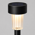 A modern solar-powered outdoor ground stake light with a matte black cylindrical top housing a dark solar panel, a ribbed clear glass shade glowing with warm light, and a black stem, shown against a neutral gray background.