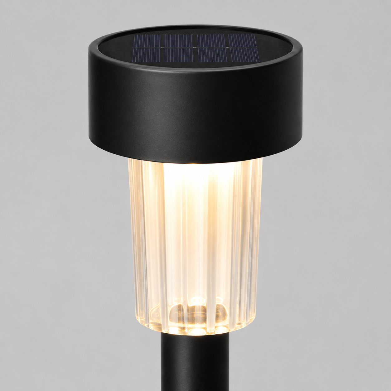 A modern solar-powered outdoor ground stake light with a matte black cylindrical top housing a dark solar panel, a ribbed clear glass shade glowing with warm light, and a black stem, shown against a neutral gray background.