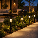Four black outdoor landscape lights with clear glass shades are installed along a garden bed next to a concrete walkway at dusk, casting a warm yellow glow. The background shows a modern patio with outdoor furniture, a stone pillar, and landscaping.