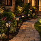 A row of outdoor solar pathway lights illuminating a stone walkway through a landscaped garden at night, with green shrubs, pink flowers, and a house in the background.