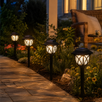 Four solar pathway landscape lights with warm white glowing glass are installed along a stone walkway in a landscaped garden bed next to a patio chair at dusk.