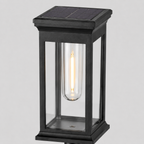 A modern solar-powered outdoor light fixture with a black metal frame, clear glass panels, and a glowing filament bulb inside, shown against a neutral light gray background.