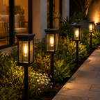 Four black outdoor solar post lanterns with warm glowing Edison bulbs, arranged along a garden bed next to a stone walkway at night.
