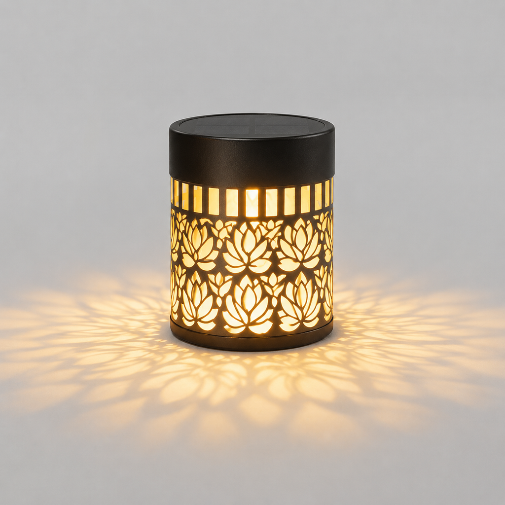 A cylindrical outdoor solar light with a black metal frame featuring a cutout lotus flower pattern, casting a warm yellow glow and shadow on a light gray background.