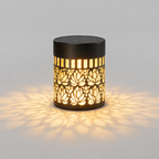 A cylindrical outdoor solar light with a black metal frame featuring a cutout lotus flower pattern, casting a warm yellow glow and shadow on a light gray background.