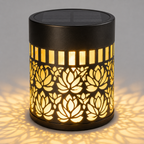 A cylindrical outdoor solar LED lamp with a black metal body, featuring a laser-cut lotus flower pattern that glows warm yellow when illuminated. The lamp is shown casting a soft shadow on a neutral gray background.