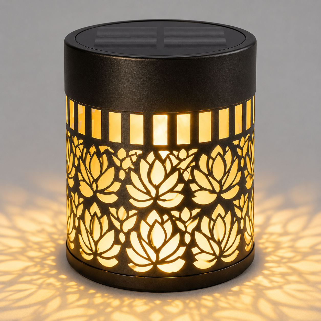 A cylindrical outdoor solar LED lamp with a black metal body, featuring a laser-cut lotus flower pattern that glows warm yellow when illuminated. The lamp is shown casting a soft shadow on a neutral gray background.