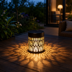 A black solar-powered outdoor lantern with intricate cutout patterns, casting a warm golden light and shadow pattern on a wooden patio deck at dusk.