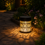 A cylindrical outdoor solar lantern with a black metal frame and glowing warm yellow light, casting a floral shadow on a stone pathway in a garden setting.