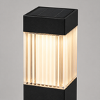 A modern solar-powered outdoor bollard post light with a black top and bottom housing and a ribbed clear glass center that glows with warm yellow light, shown against a neutral gray background.