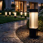 A modern outdoor landscape light with a black rectangular base and a ribbed clear glass cylinder, glowing with warm light, situated on a pebble bed next to a stone walkway in a landscaped garden at night.