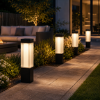 Four modern outdoor solar pathway lights with black bases and glowing white ribbed glass panels, arranged along a stone walkway in a landscaped garden at night.