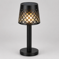 A black table lamp with a cylindrical base and a perforated shade that glows with warm light, shown on a neutral gray background.