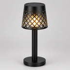 A black table lamp with a cylindrical base and a perforated shade that glows with warm light, shown on a neutral gray background.