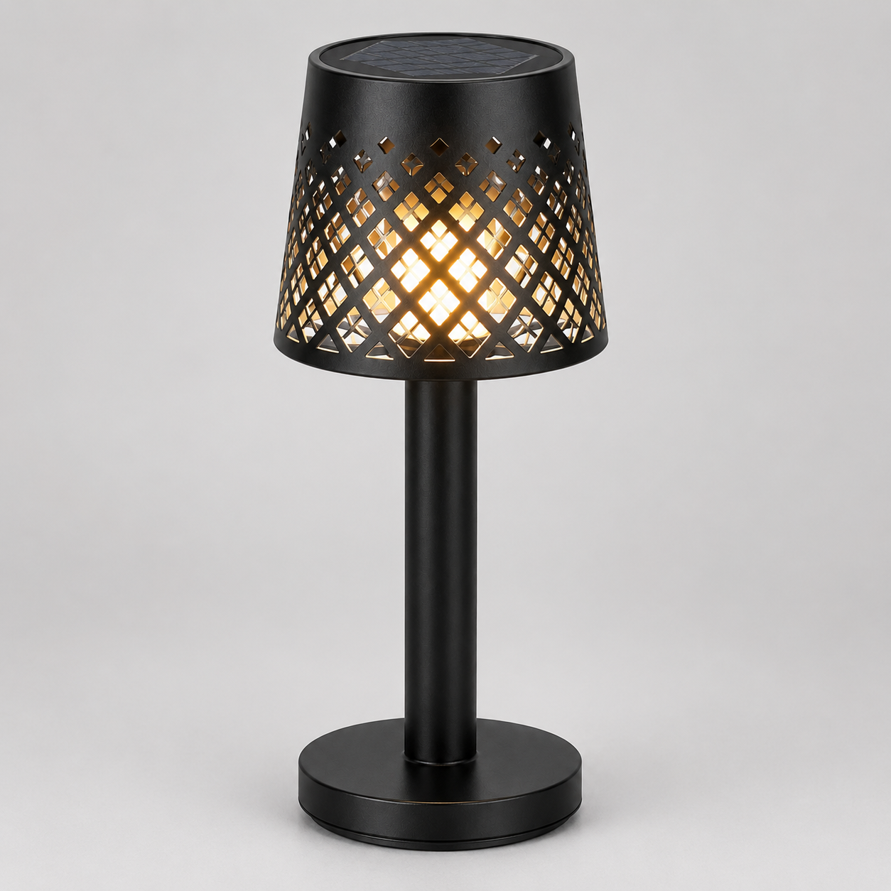 A black table lamp with a cylindrical base and a perforated shade that glows with warm light, shown on a neutral gray background.