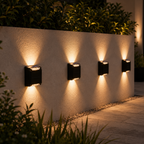 Four black square outdoor wall sconces mounted on a textured beige wall at night, casting warm light downward in a garden setting with plants and a paved walkway.