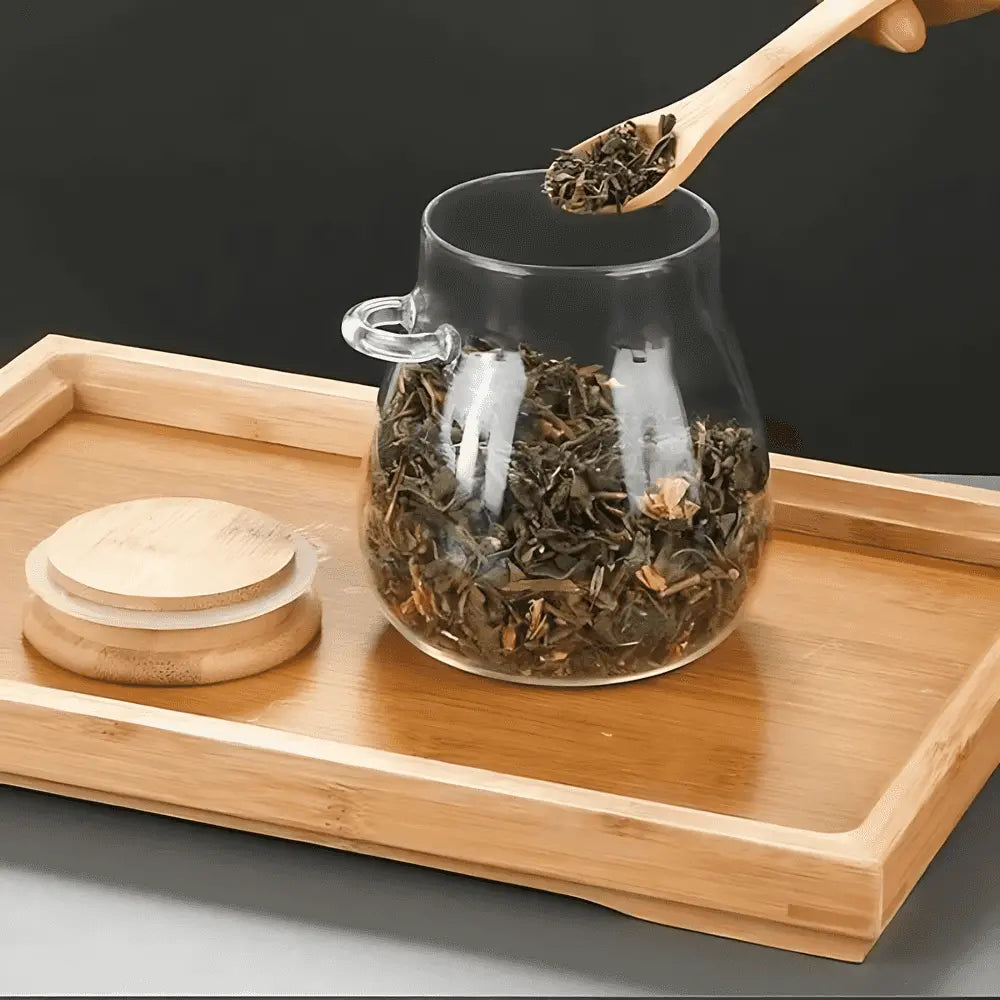 Bamboo spoon lifting loose tea from an open glass storage jar