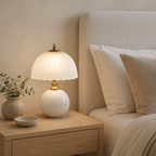 A white half dome table lamp glows beside an upholstered cream bed.
