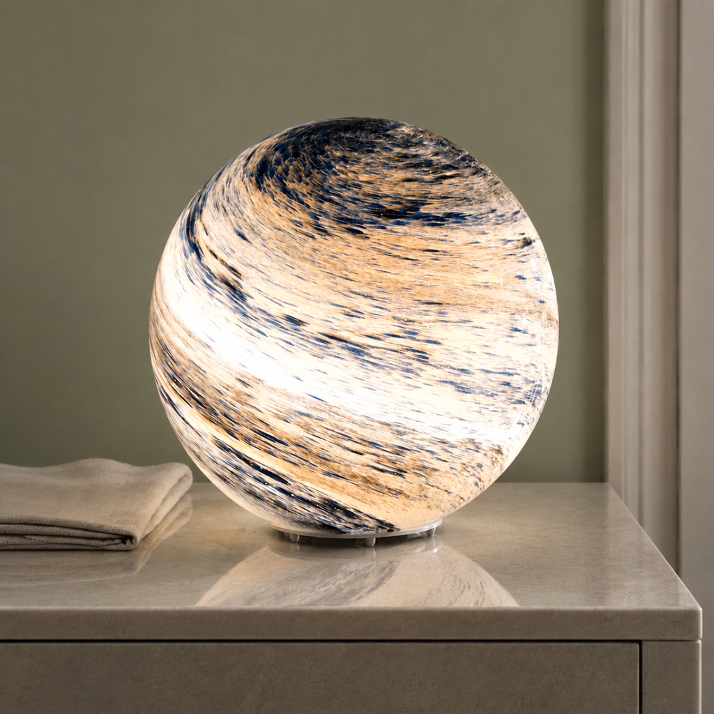 Glass moon table lamp glowing warm white on a stone bedside table beside folded linen.