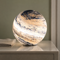 Glass moon table lamp glowing warm white on a stone bedside table beside folded linen.