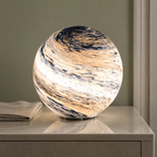 Glass moon table lamp glowing warm white on a stone bedside table beside folded linen.