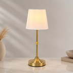 Gold cordless table lamp with white fabric shade on a light gray background.