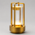 Gold cordless lantern table lamp with a clear cylindrical shade, illuminated against a light gray background.