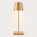 Gold table lamp with a tapered shade, slim stem, square base and warm light on a plain background.