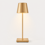 Gold table lamp with a tapered shade, slim stem, square base and warm light on a plain background.
