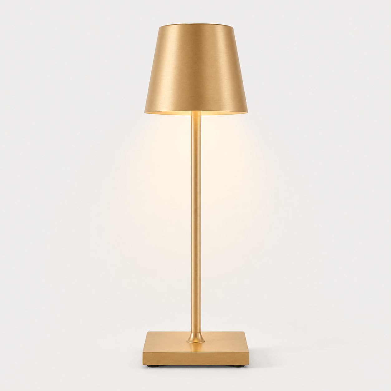 Gold table lamp with a tapered shade, slim stem, square base and warm light on a plain background.