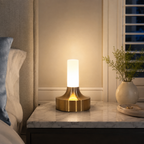 Gold table lamp glowing on a bedside table beside pillows and curtains.