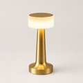 Gold table lamp with a glowing white cylindrical shade on a plain light gray background.