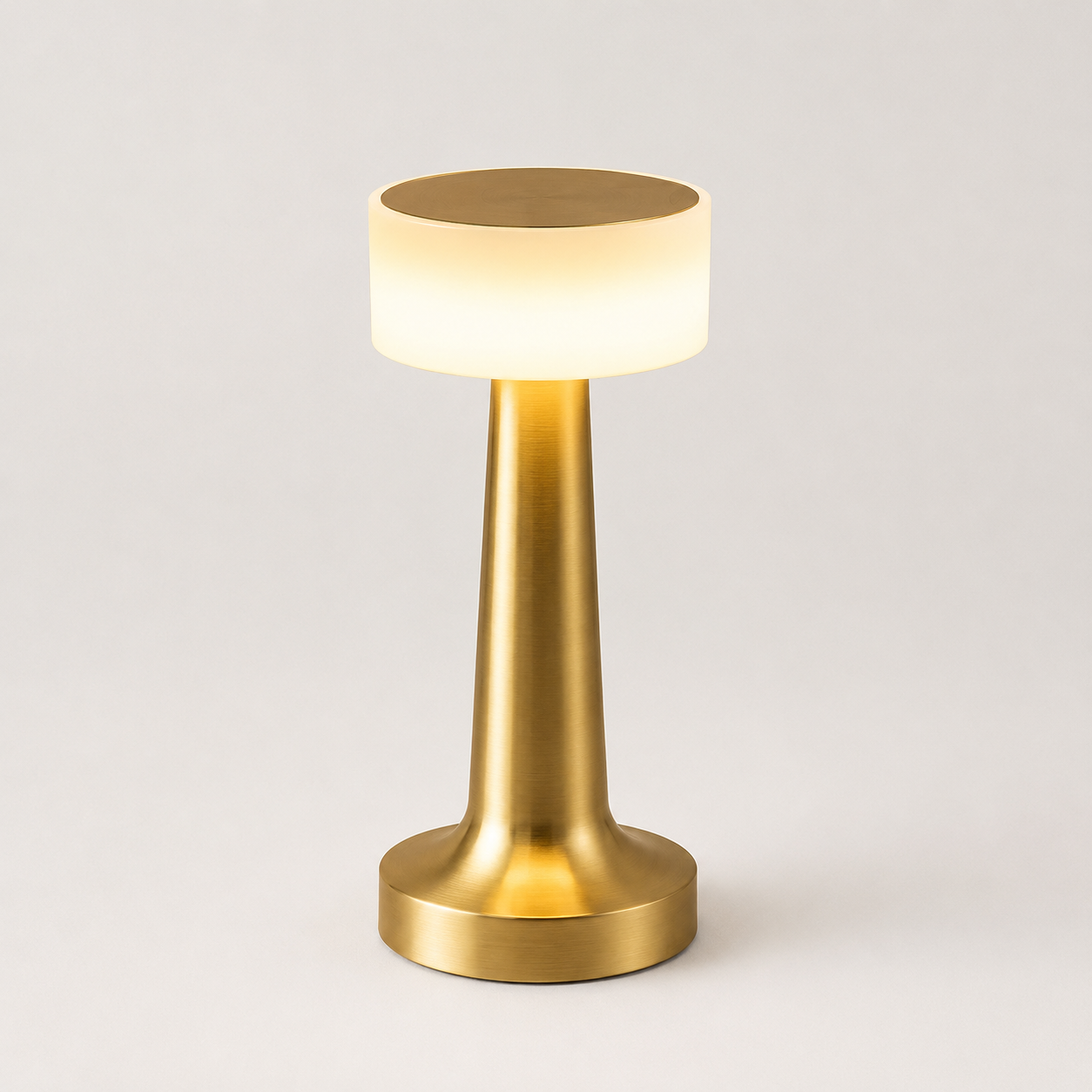 Gold table lamp with a glowing white cylindrical shade on a plain light gray background.