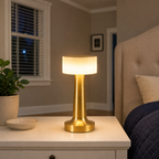 Gold table lamp lit on a white bedside table beside a bed and window.