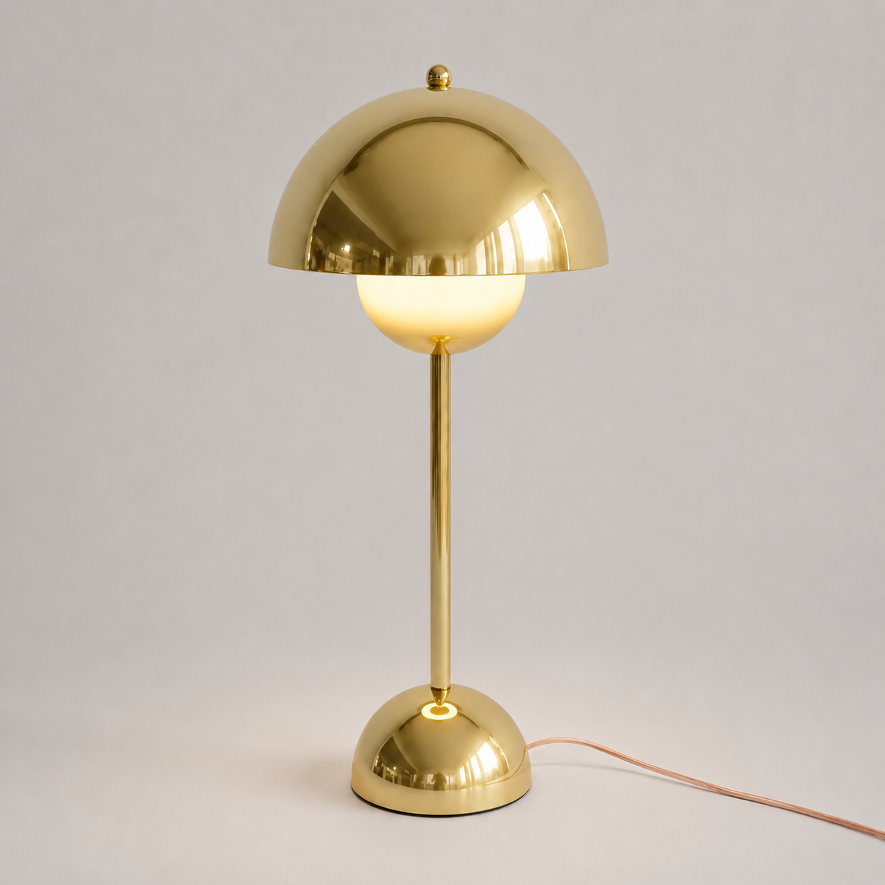 Gold table lamp with a reflective dome shade, slim stem, circular base, power cord, and warm downward glow.
