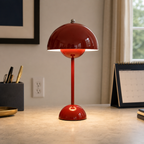 Red table lamp on a light desk near framed artwork, stationery, and a bright window.
