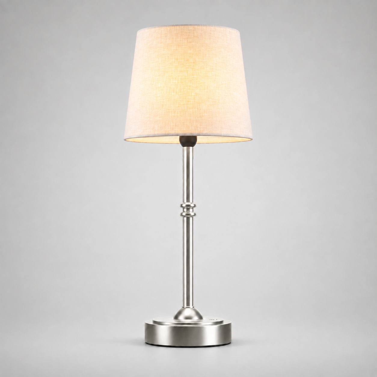 Silver table lamp with a white fabric shade, illuminated against a light gray background.