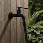 Wall-mounted faucet in matte black mounted on wood with a garden hose attached.